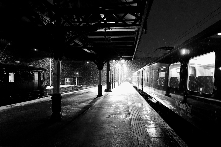 Black and white photo showing a train station platform at night with heavy rain and bright lights
