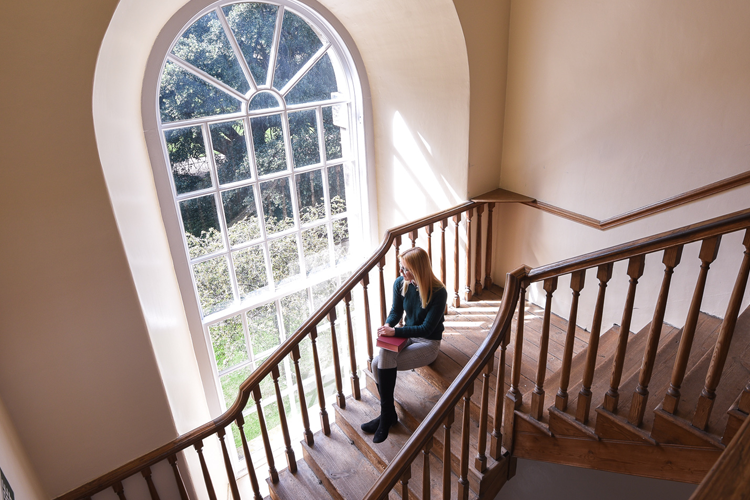 Person sits on a wooden staircase and looks out of a large arched window.