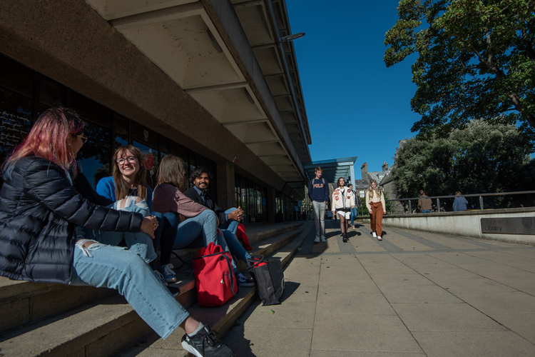 Students sit on steps. Other students walk towards them down a paved walkway.