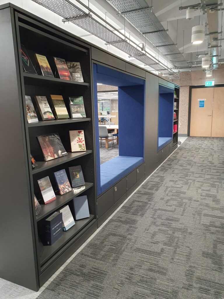 A black shelving unit of two bookcases containing books, between the two bookcases are blue cushioned seating areas. In the background the door to the Postgraduate area on level 1 can be seen.