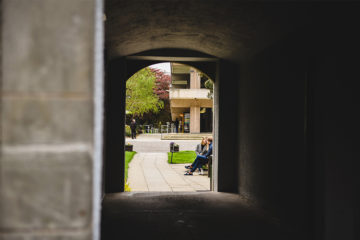 Main Library seen through archway approaching entrnace. two people sit on a bench outside