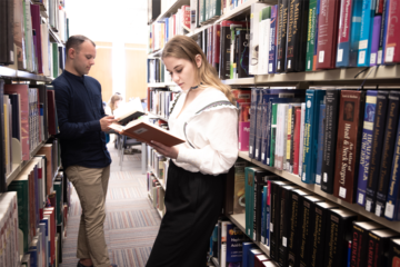 Two students stand at bookshelves. Both are reading books.