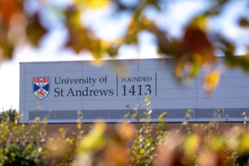 University of St Andrews crest is displayed on a large grey building, the image is framed by slightly out of focus autumn leaves.
